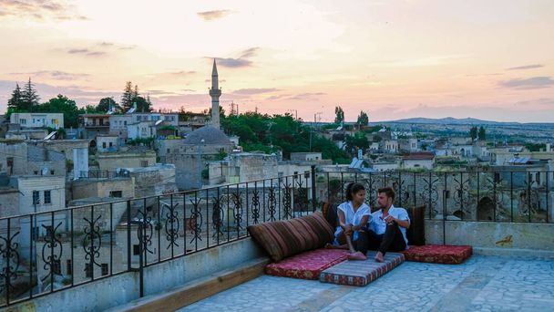 happy couple with a drink on the rooftop of a cave house historical village of Ayvali Goreme Cappadocia, couple watch sunset Kapadokya.  - Foto, imagen