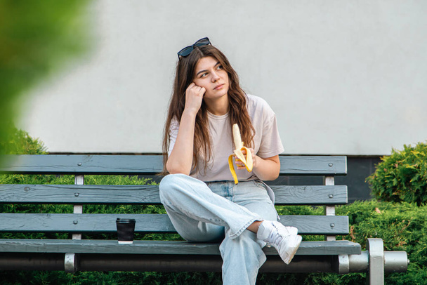 Eine traurige junge Frau sitzt auf einer Bank und isst Banane, Snack-Konzept. - Foto, Bild