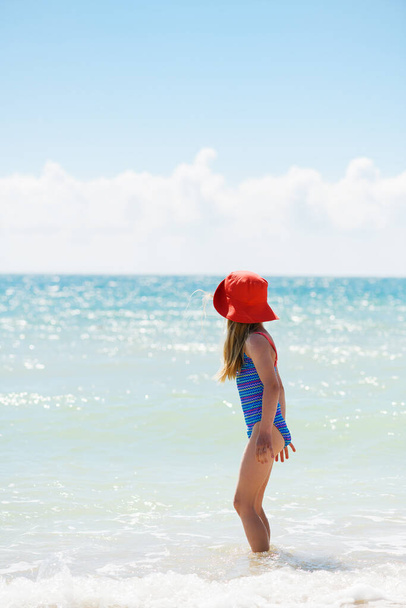 Little girl child stay in water in swimsuit and sun hat, look back at beautiful sea horizon, happy family vacation, vertical shot - Photo, Image