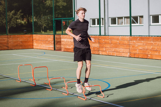 Blond boy in sportswear jumps over red hurdles to improve lower body dynamics. Plyometric training in an outdoor environment. Improve your skills. - Zdjęcie, obraz