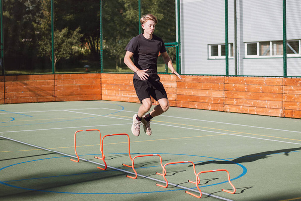 Blond jongetje in sportkleding springt over rode obstakels om de dynamiek van het onderlichaam te verbeteren. Plyometrische training in een buitenomgeving. Verbeter uw vaardigheden. - Foto, afbeelding
