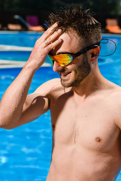 Smiling young man portrait in swimming goggles in pool, summer vacation activity, fit sportsman in polarized glasses - Photo, Image
