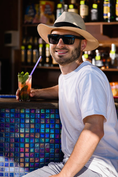 Mladý usměvavý muž v slunečních brýlích a slamáku pije koktejl v beach baru, letní prázdniny vibruje - Fotografie, Obrázek