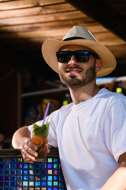 Nuori hymyilevä mies aurinkolaseissa ja olkihattu juominen cocktail rantabaarissa, kesäloma vibes - Valokuva, kuva