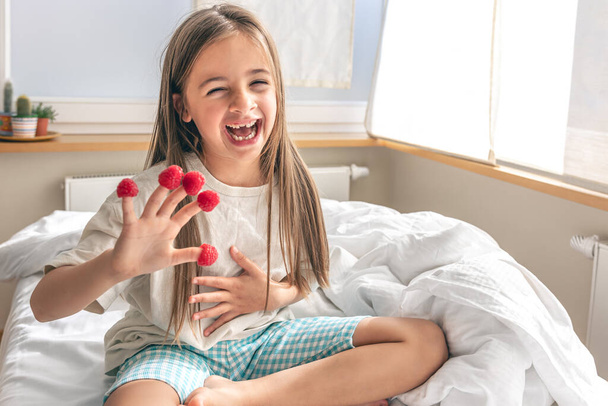 Drôle de petite fille avec des framboises sur ses doigts se trouve dans le lit le matin, copier l'espace. - Photo, image