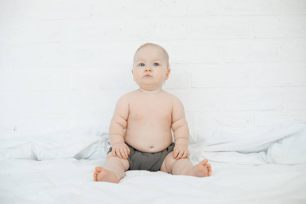 Hermoso bebé de ojos azules se sientan en una sábana blanca en la cama y miran hacia arriba, espacio de copia gratuito. Retrato de un encantador bebé desnudo sobre fondo blanco. Concepto de infancia feliz y cuidado de niños. - Foto, imagen