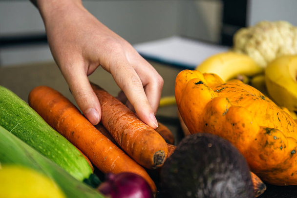 Zbliżenie, ręka kobiety bierze marchewkę wśród warzyw na stole kuchennym do gotowania. - Zdjęcie, obraz