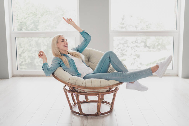 Photo of excited charming young girl wear jeans shirt sitting chair smiling having rest pointing finger indoors house room. - Photo, Image