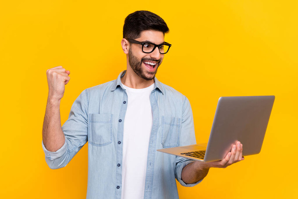 Portrait of young handsome guy holding laptop fist up celebrates good marks online education isolated on yellow color background. - Foto, immagini