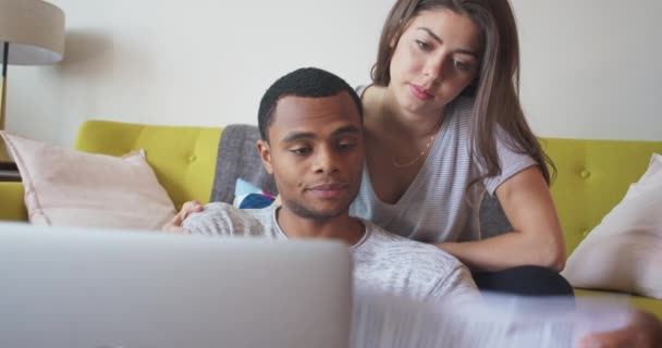 Afrikai-amerikai és kaukázusi pár online fizet számlákat laptoppal. Millenniumi friss házasok ülnek a nappaliban, és a papírokat nézegetik. 4k Lassú mozgás kézi - Felvétel, videó