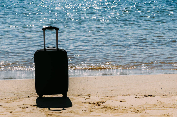 Μαύρη βαλίτσα ταξιδιού σε αμμώδη παραλία με τυρκουάζ φόντο στη θάλασσα, καλοκαιρινές διακοπές έννοια - Φωτογραφία, εικόνα