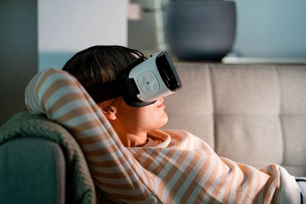 Happy woman lying on cozy sofa resting after hardworking day. Female wearing VR glasses plays interesting games with amazing graphics and explores virtual reality at home at night. Cyberspace concept - Foto, imagen