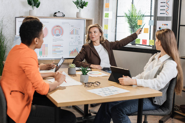 Caucasian mature woman in suit showing financial statistic of enterprise on glass board during working meeting with diverse females colleagues. Cooperation, technology and people concept. - Photo, Image