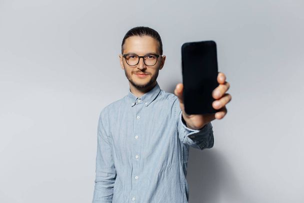 Studio portrait of young smiling man showing a smartphone on light grey background. - Fotoğraf, Görsel