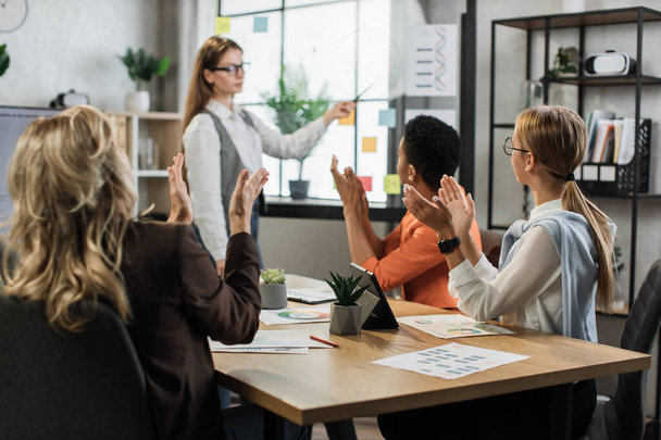 Caucasian woman pointing glass board with financial report during conference with multiracial female partners. Business woman colleagues gathering at office room for brainstorming. - Foto, imagen