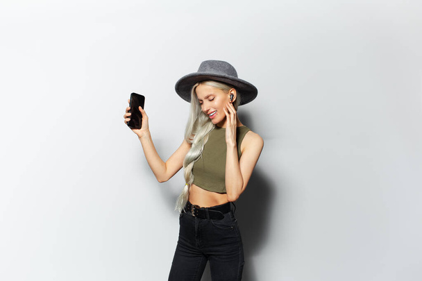 Studio portrait of beautiful young happiness blonde girl while dancing and listen the music via wireless earbuds, holding smartphone in hand, wearing grey hat on white background. - Фото, зображення
