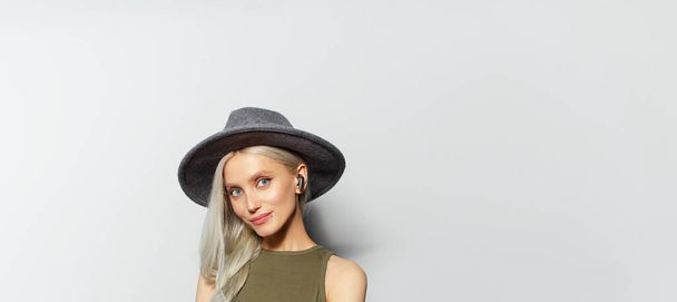 Studio portrait of young pretty blonde girl with wireless earbuds in ear, wearing grey hat on white background. Panoramic banner with copy space. - Photo, Image