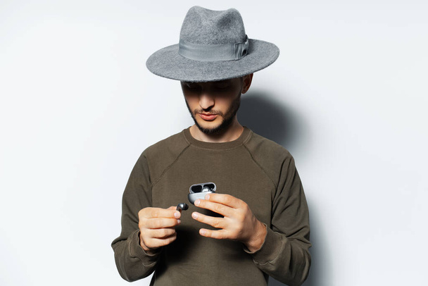 Studio portrait of young man holding wireless earbuds and charger case in hands on white background, wearing sweater and grey hat. - Photo, image