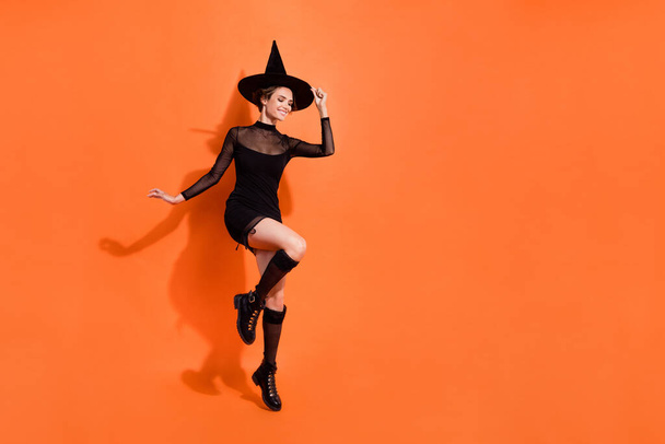 Full body photo of attractive young woman jump touch hat smile wear stylish black halloween witch look isolated on orange color background. - Fotoğraf, Görsel