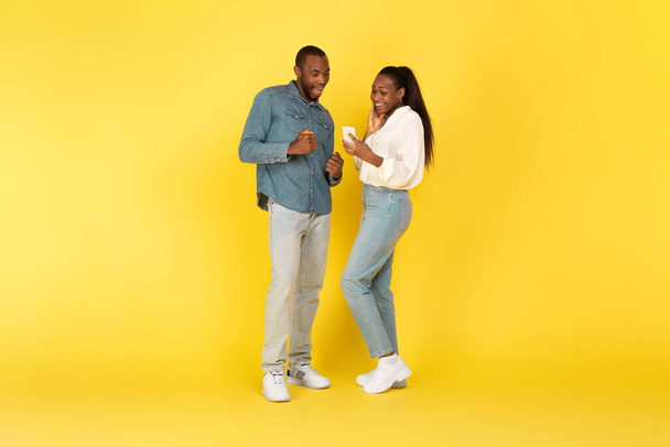 黄色のスタジオの背景に立つ携帯電話のテキストとウェブサーフィンを使用して興奮したアフリカ系アメリカ人のカップル。携帯電話を示す妻と夫への偉大なアプリケーション.全長 - 写真・画像