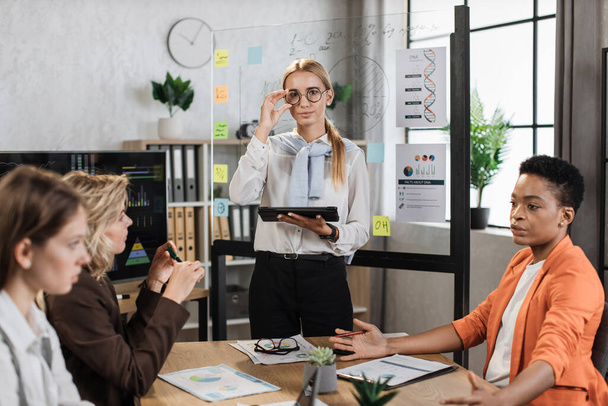 Portrait of confident blond woman in white shirt and eyewear posing at modern office. Three diverse female coworkers talking sitting near their leader at office. - Fotografie, Obrázek