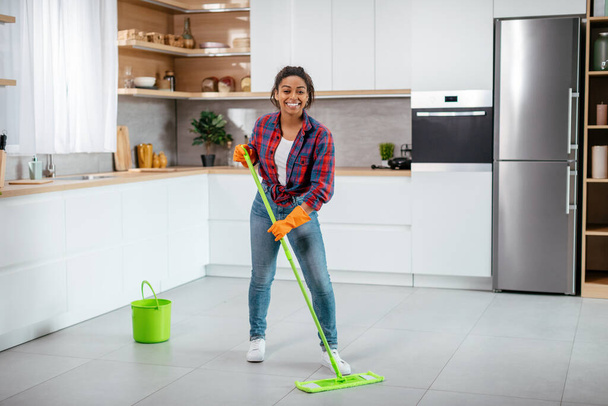 Happy young black woman in rubber gloves washes floor with mop in minimalist kitchen interior, full length, copy space. Household chores, cleanliness and hygiene, health care and house cleaning alone - Fotó, kép