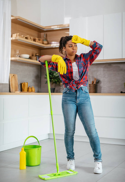 Unhappy tired millennial black female in rubber gloves washes floor with mop, wiping sweat from forehead in kitchen interior. Overwork from household chores, cleanliness and hygiene at home alone - Photo, Image