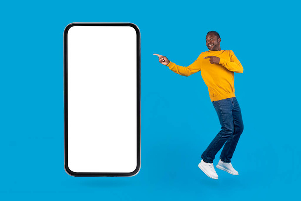 Concepto de aplicación móvil. Alegre chico afroamericano bailando por teléfono celular grande sobre fondo de estudio azul, hombre afroamericano apuntando a teléfono inteligente con pantalla vacía blanca, maqueta, tiro de cuerpo entero - Foto, imagen
