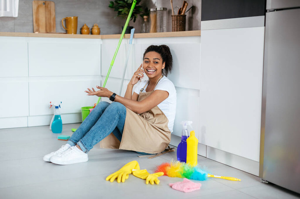 Happy millennial black woman sits on floor with mop, cleaning supplies, calls by phone in kitchen interior. Gossip, break from a lot of household chores, cleanliness and hygiene at home in free time - Photo, image