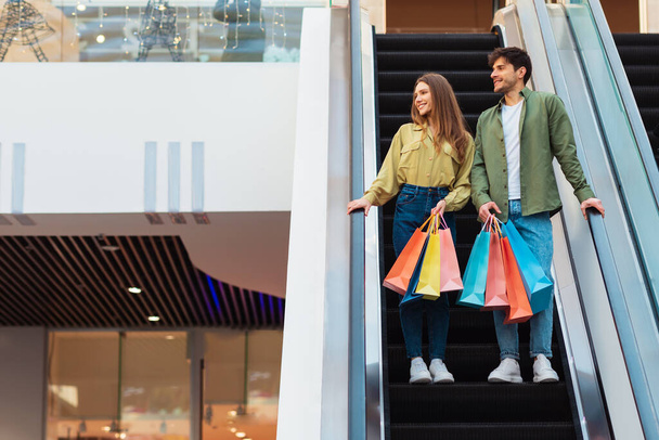 Einkaufen und Verkaufen. Junge Ehepartner stehen auf beweglichen Treppen und halten Einkaufstaschen in der Hand, um in modernen Hypermarket-Innenräumen neue Kleidung zu kaufen. Paar verbringt Wochenende in Einkaufszentrum. Volle Länge - Foto, Bild