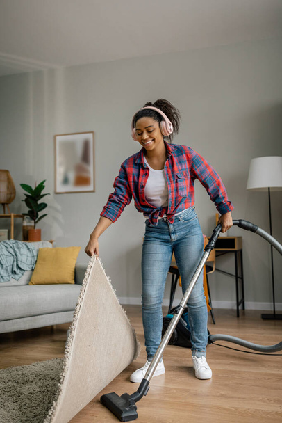 Cheerful young black female in wireless headphones with vacuum cleaner dusts floor, enjoys music in living room interior. Audio app and fun, cleaning housework, cleanliness and hygiene at home alone - Photo, image