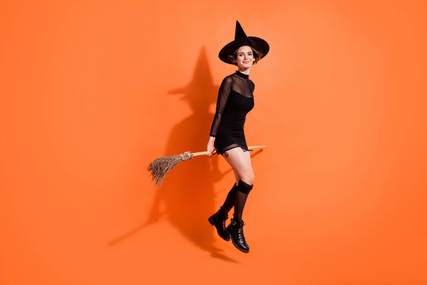 Full length photo of charming young lady sit broomstick soar wear stylish black halloween witch clothes isolated on orange color background. - Fotó, kép