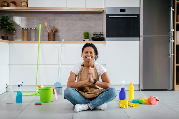 Glad young black lady sit on floor with mop, cleaning supplies meditating in white kitchen interior. Peace and calm, household chores, cleanliness and hygiene, health care alone at home in free time - Photo, Image