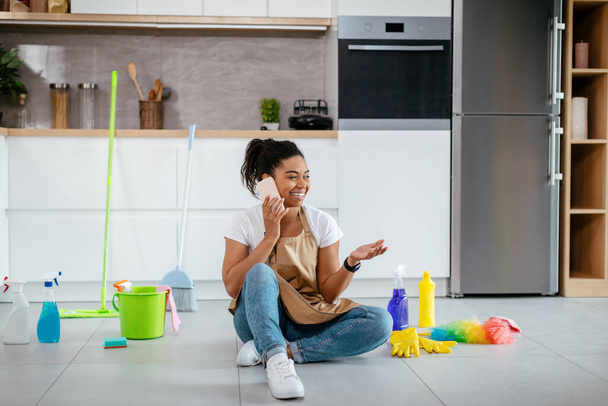 Χαίρομαι νεαρή μαύρη γυναίκα κάθεται στο πάτωμα με προμήθειες καθαρισμού κλήσεις μέσω τηλεφώνου στο σύγχρονο εσωτερικό της κουζίνας. Νέα, κουτσομπολιά, καλή προσφορά και διάλειμμα από τις δουλειές του σπιτιού, καθάρισμα μόνος στο σπίτι το Σαββατοκύριακο - Φωτογραφία, εικόνα