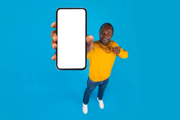 Felice bello afroamericano uomo di mezza età in casual mostrando nuovissimo cellulare con schermo bianco vuoto, sfondo blu studio, panorama con spazio copia, mockup, vista ad alto angolo, lunghezza intera - Foto, immagini