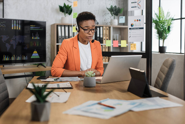 Schöne afrikanisch-amerikanische Frau in formeller Kleidung und Headset sitzt am Schreibtisch und tippt auf einem drahtlosen Laptop. Weibliche Person mit kurzen Haaren arbeitet im Büro auf tragbarem Computer. - Foto, Bild