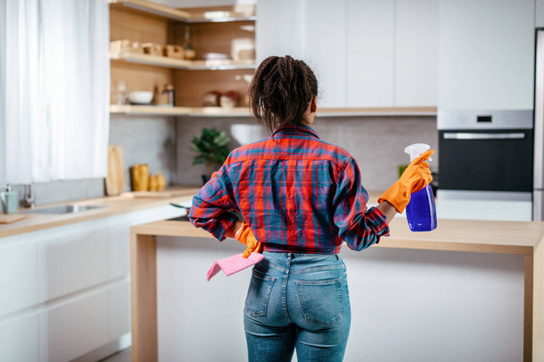 Drukke jonge Afrikaanse Amerikaanse dame in rubberen handschoenen met spray, spons kijkt naar schoonmaak resultaat in minimalistische keuken interieur, rug, kopieerruimte. Huishoudelijke taken, reinheid en hygiëne, gezondheidszorg - Foto, afbeelding