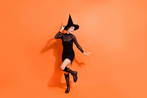 Фотографія повного тіла чарівної молодої дівчини, танцюючої костюмованої вечірки, одягнена в стильний чорний одяг відьми Хеллоуїна ізольована на помаранчевому кольорі
. - Фото, зображення