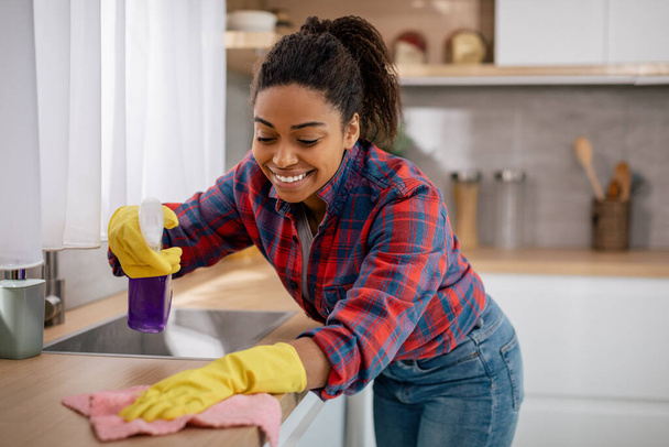 Vrolijke jonge zwarte vrouw veegt tafel van vuil met schoonmaakmiddelen in minimalistische keuken interieur. Lifestyle, schoonmaak en huishoudelijke taken, netheid en hygiëne, gezondheidszorg alleen thuis - Foto, afbeelding