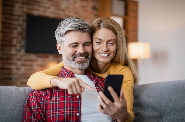 Щаслива жінка середнього віку, яка обіймає чоловіка з смартфоном, має відео-дзвінок, сидить на дивані в затишній вітальні. Пристрій для зустрічі віддалено, додаток для комунікації, реклама і пропозиція належної covid-19 - Фото, зображення