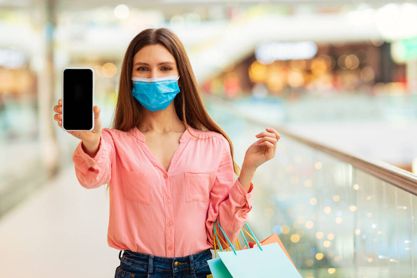 Mobilne zakupy. Lady Showing Cellphone With Blank Screen To Camera Pozowanie noszenie maski ochronnej twarzy stojącej w hipermarkecie w pomieszczeniach. Koncepcja zastosowania w handlu elektronicznym - Zdjęcie, obraz