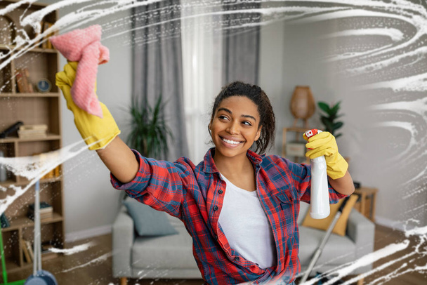 Glad young black lady in rubber gloves washes mirror with spray, enjoys cleaning in living room interior. Great work, household chores, cleanliness and hygiene at home alone, lifestyle and emotions - Photo, Image