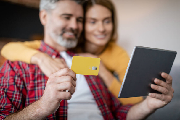 Nakupování online doma. Šťastný dospělý Evropan dáma objetí muž s kreditní kartou a tablet v obývacím pokoji interiéru, rozmazané, zblízka. Koupit objednávku, prodej pro shopaholics, skvělá nabídka a reklama due covid-19 - Fotografie, Obrázek