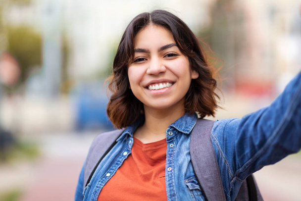 Fröhliche junge Frau aus dem Nahen Osten beim Selfie im Freien auf der City Street, glückliche arabische Studentin mit Rucksack, die ein Selbstporträt aufnimmt und in die Kamera lächelt, während sie draußen steht - Foto, Bild