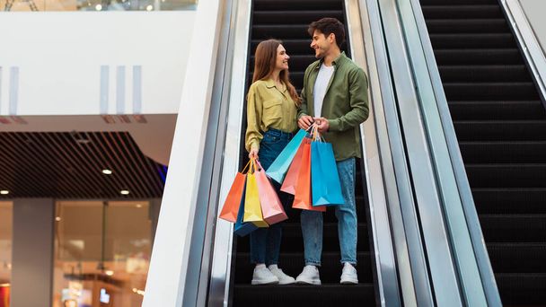 Millennial ζευγάρι ψώνια μαζί κρατώντας τσάντες Shopper στέκεται στις κινούμενες σκάλες στο σύγχρονο εμπορικό κέντρο εσωτερικούς χώρους. Νεαρές σύζυγοι που αγοράζουν νέα ρούχα ξοδεύουν το Σαββατοκύριακο στο Hypermarket. Πανόραμα - Φωτογραφία, εικόνα