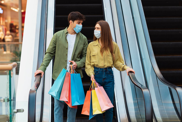 Başarılı bir alışveriş. Alışveriş merkezlerinde maske takan ve hareket halindeki merdivenlerde duran çift. Eşler hafta sonları modern hipermarkette vakit geçiriyorlar. Satış Sezonu Konsepti - Fotoğraf, Görsel