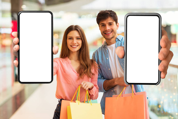 Shopping-App. Paar zeigt riesige Mobiltelefone mit leeren Bildschirmen, die im modernen Hypermarkt stehen und Einkaufstaschen halten. Ehepartner, die den Kunden eine Antragsempfehlung geben. Attrappe - Foto, Bild