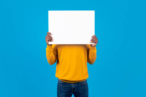 Unrecognizable african american guy in casual outfit hiding his face behind empty white advertising board, blue studio background, copy space. Human emotions, thoughts concept - Foto, immagini
