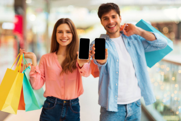 Coppia allegra che mostra lo schermo dello smartphone raccomanda un'applicazione di shopping mobile in posa con borse colorate nell'ipermercato moderno. Concentrazione selettiva sui telefoni. Mockup - Foto, immagini