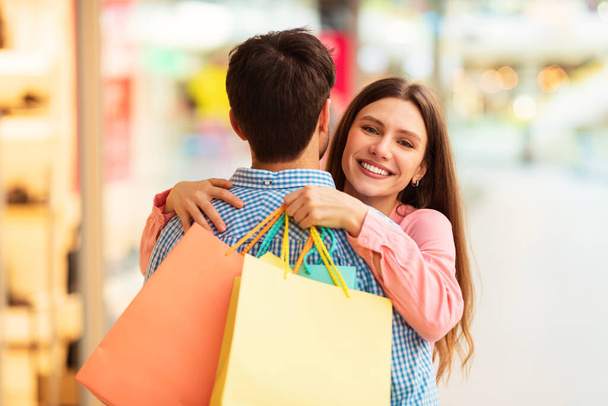 Ευτυχισμένη σύζυγος αγκαλιάζει τον σύζυγο απολαμβάνοντας ψώνια μαζί στο σύγχρονο εμπορικό κέντρο το Σαββατοκύριακο. Σύζυγοι Ποζάρουν Αγκαλιάζοντας Κρατώντας Τσάντες Shopper Σε Υπεραγορά. Μεγάλη έννοια προσφοράς πωλήσεων. Επιλογή εστίασης - Φωτογραφία, εικόνα